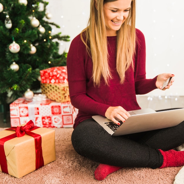 Smiling lady with laptop and plastic card near gift boxes and Christmas tree