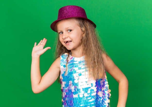 smiling little blonde girl with purple party hat standing with raised hand isolated on green wall with copy space