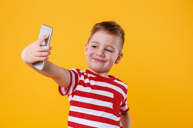 Smiling little boy holding mobile phone and making selfie