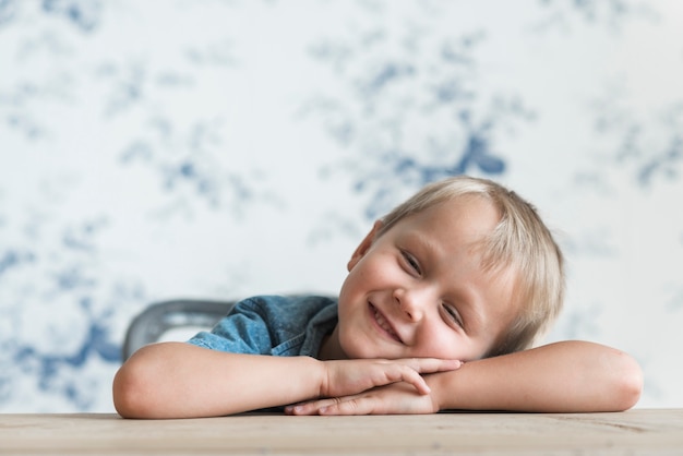 Free photo smiling little boy leaning his head on hand over the wooden table