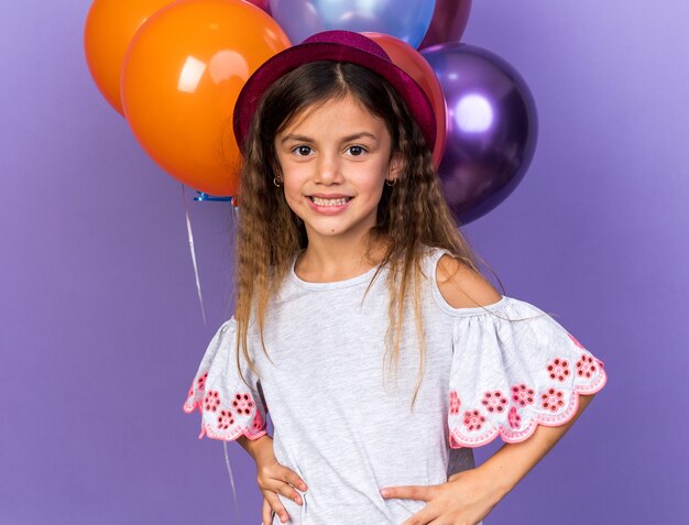 smiling little caucasian girl with violet party hat standing in front of helium balloons isolated on purple wall with copy space