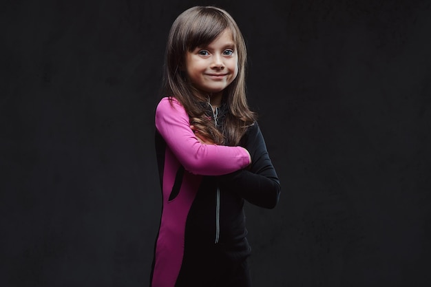 Smiling little girl dressed in sportswear standing with crossed arms. Isolated on a dark textured background.