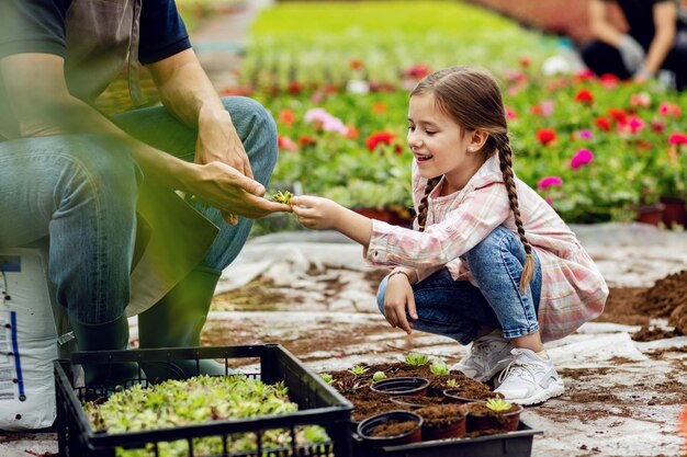 Smiling little girl having fun while assisting her father and planting flowers in a pot at plant nursery