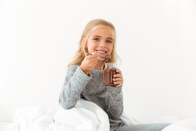 Free photo smiling little girl holding bank of sweet chocolate hazelnut spread,  while sitting in bed