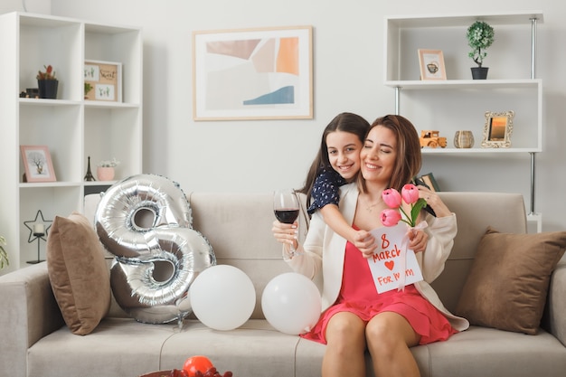 Smiling little girl standing behind sofa holding flowers with greeting card hugged mother on sofa on happy women's day in living room