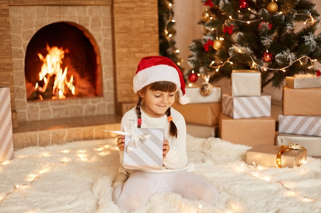 Smiling little girl wearing white sweater and santa claus hat, sitting on floor near Christmas tree, present boxes and fireplace, holding present from parents in hands.