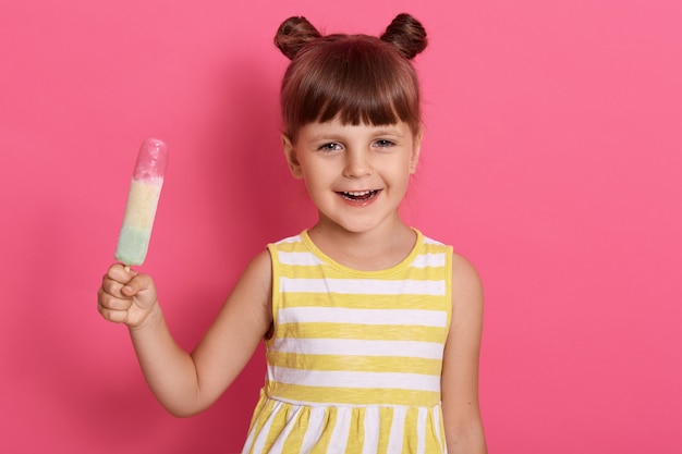 Smiling little girl with ice cream posing isolated over rose wall with joyful expression, holding water ice in hands, funny female kid with knots, wearing summer dress.