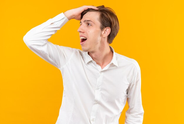 Smiling looking side young handsome guy wearing white shirt putting hand on head isolated on orange wall