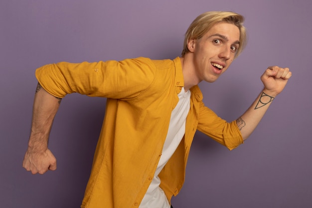 Smiling looking straight ahead syoung blonde guy wearing yellow t-shirt showing running gesture isolated on purple