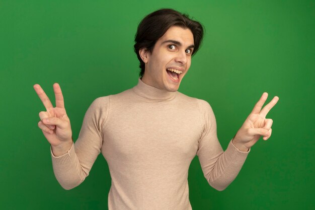 Smiling looking  young handsome guy showing peace gesture isolated on green wall