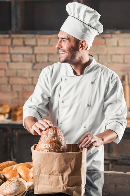 Free photo smiling male baker putting bread in brown paper bag