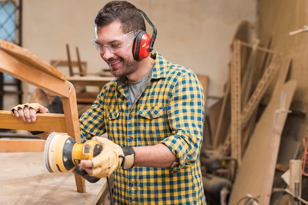 Free photo smiling male carpenter sanding a wood with orbital sander in workbench