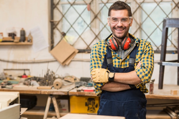 Free photo smiling male carpenter wearing safety glasses standing in front of workbench with his arm crossed