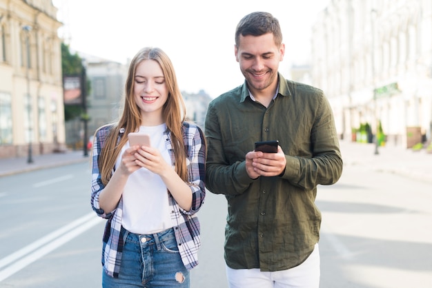 Free photo smiling male and female friends using cellphone white walking together on street