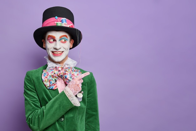 Free Photo smiling man dresses for carnival party has image of hatter from wonderland indicates away on blank space wears halloween costume and bright makeup isolated on purple wall