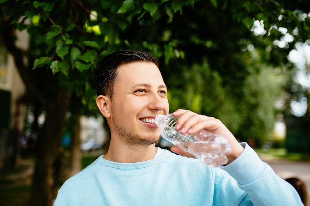 Smiling man drinks water from bottle on the background of green tree in summer day