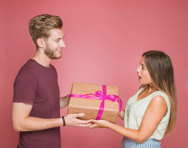 Smiling man giving gift box to her shocked girlfriend against colored background