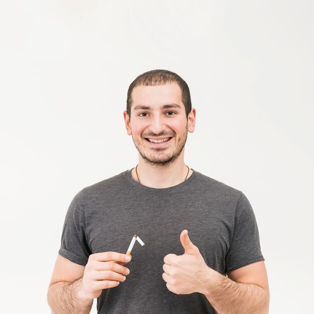 Smiling man holding broken cigarette showing thumb up sign against white background