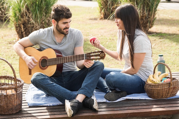 Smiling man playing guitar with his girlfriend holding red apple in hand