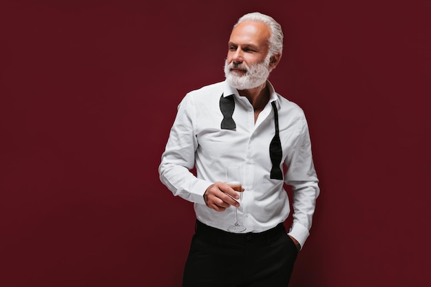 Free photo smiling man in suit holds champagne glass fashionable guy with grey beard in classic outfit posing on burgundy background