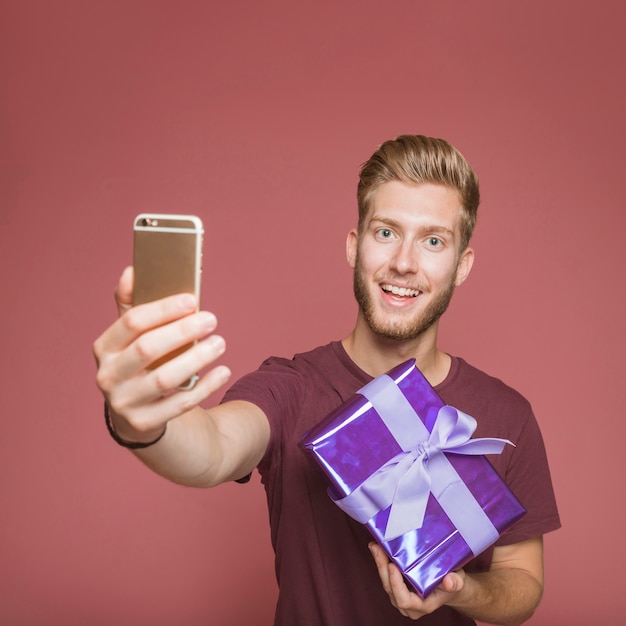 Free photo smiling man taking self portrait from mobile holding gift box