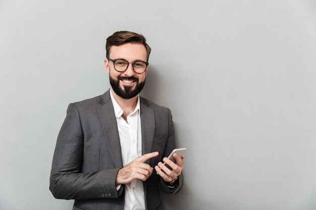 Smiling man in white shirt typing text message or scrolling feed in social network using smartphone over gray