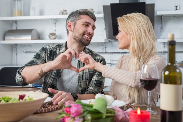 Smiling man and woman showing heart by hands and sitting at table in kitchen