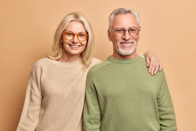 Smiling mature couple embrace look gladfully at camera pose for family portrait happy children came to visit them wear transparent glasses casual jumpers isolated over brown wall