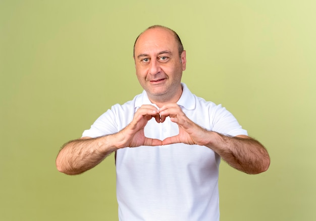 Smiling mature man showing heart gesture isolated on olive green wall