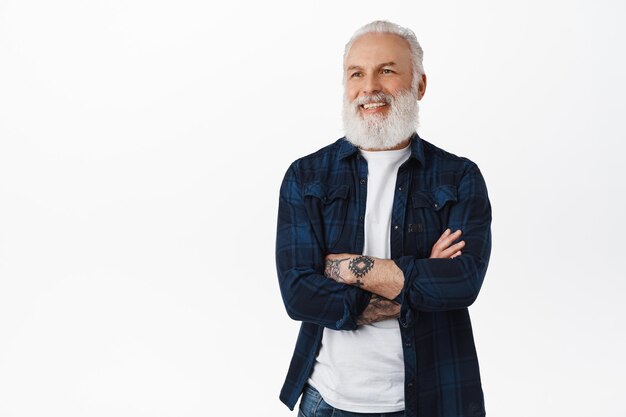 Smiling mature senior man with tattoos and grey long beard, cross arms on chest confident, looking left at promotional text logo, pleased to see advertisement, standing over white background