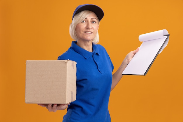 Free photo smiling middle-aged blonde delivery woman in blue uniform and cap holding cardbox and clipboard looking at camera isolated on orange background