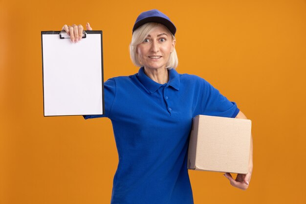 smiling middle-aged blonde delivery woman in blue uniform and cap holding cardbox and showing clipboard  isolated on orange wall