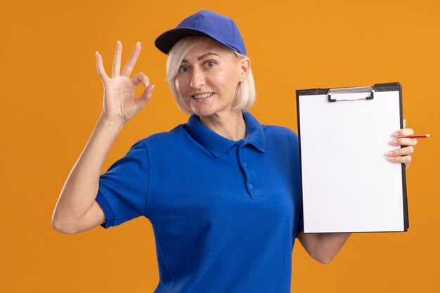 Smiling middle-aged blonde delivery woman in blue uniform and cap showing clipboard to camera  doing ok sign isolated on orange wall