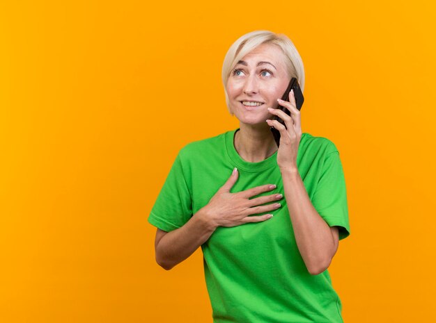 Smiling middle-aged blonde slavic woman talking on phone keeping hand on chest looking at side isolated on yellow wall with copy space