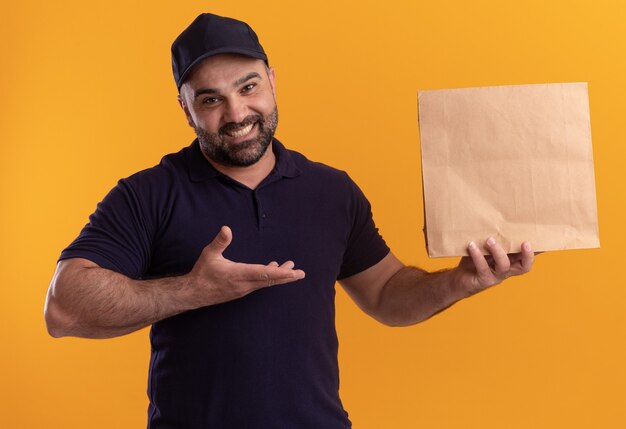 Smiling middle-aged delivery man in uniform and cap holding and points at paper food package isolated on yellow wall