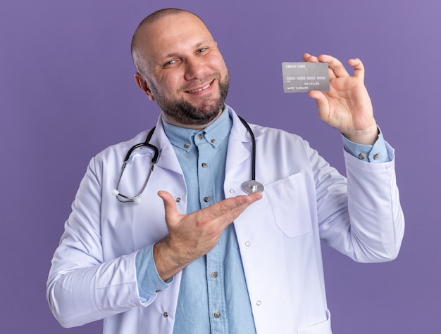 Smiling middle-aged male doctor wearing medical robe and stethoscope holding and pointing at credit card  isolated on purple wall