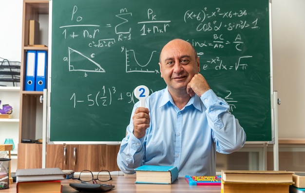 Free Photo smiling middle-aged male teacher sits at table with school supplies holding number fans in classroom