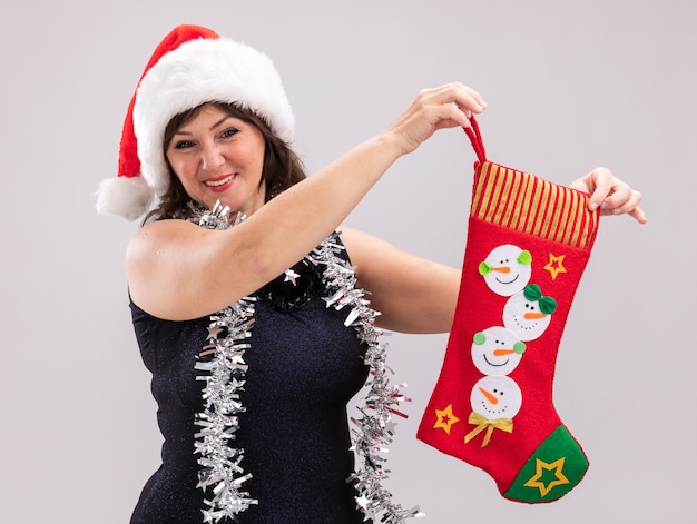 Free photo smiling middle-aged woman wearing santa hat and tinsel garland around neck holding christmas stocking looking at camera isolated on white background