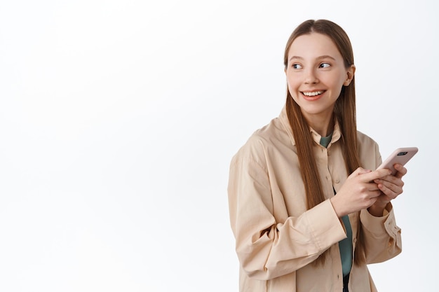 Free Photo smiling millennial girl chatting on social media, using app on smartphone, turn head at logo, looking behind with pleased smile, standing over white background. networking and technology concept