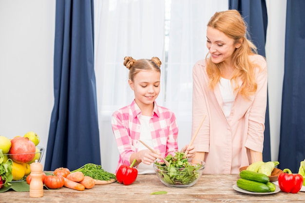 Free photo smiling mother looking at her daughter preparing salad in the glass bowl on wooden table