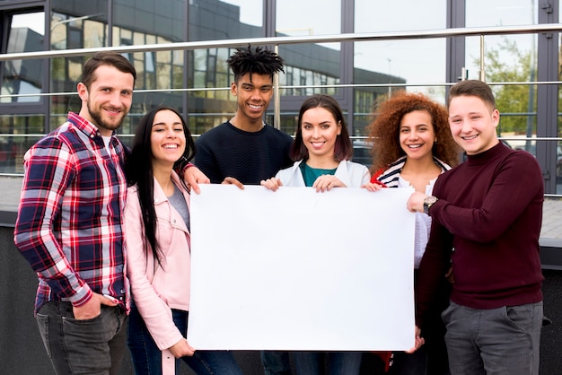 Smiling multiethnic students holding blank white poster standing in front of glass building