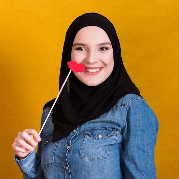 Free photo smiling muslim woman holding a paper prop in the shape of red lips over backdrop