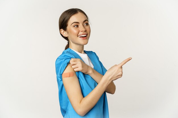 Smiling nurse female healthcare worker in scrubs pointing finger right showing patch on shoulder vac...