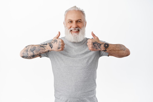 Free photo smiling old man shows thumbs up and looks happy and proud, praise great work, excellent job, standing satisfied and pleased against white wall