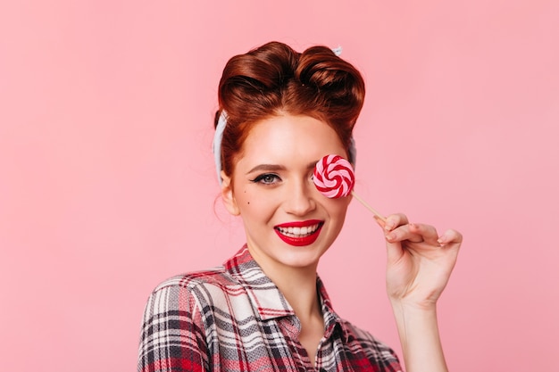 Smiling pinup girl eating red lollipop. Front view of woman in checkered shirt isolated on pink space.