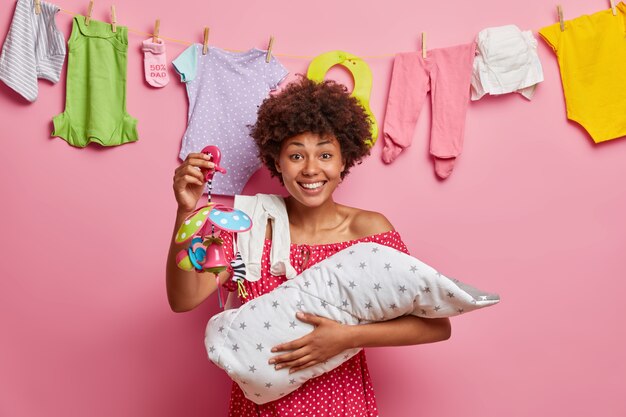 Smiling pleased ethnic mother shows mobile toy to her small infant, plays with newborn son, happy to become mom, stands indoor against pink wall. Baby in mums arms. Child care concept.