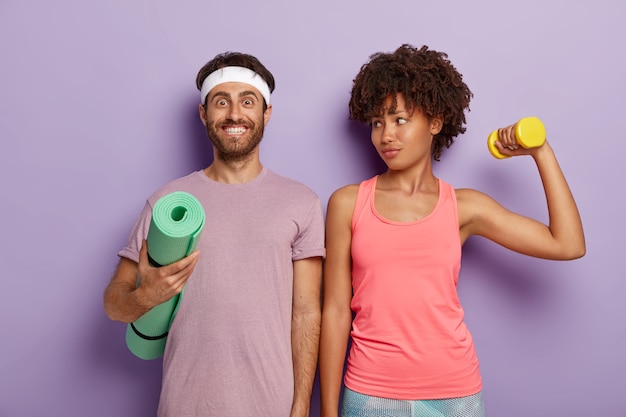 Free photo smiling pleased man poses with fitness mat, wears purple t shirt and headband, lovely sporty woman looks at husband, trains biceps with weight, stand shoulder to shoulder indoor. aerobics and people