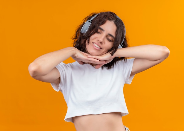 Smiling pleased young beautiful woman wearing headphones and putting hands under her chin on isolated orange wall