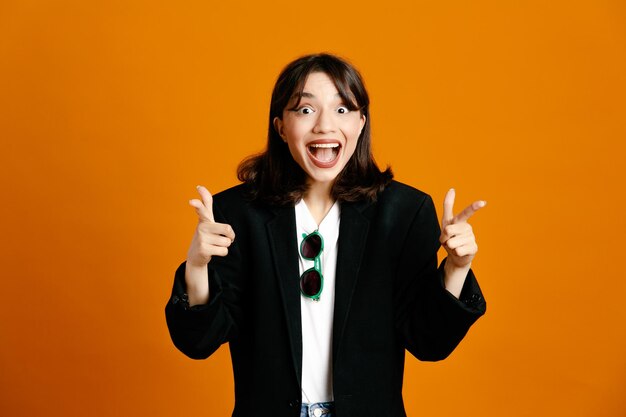 Smiling points at camera young beautiful female wearing black jacket isolated on orange background