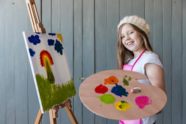 Smiling portrait of a blonde girl holding wooden palette in hand painting the canvas